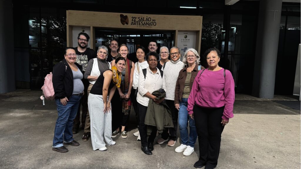 Na imagem aparece um grupo de pessoas olhando para tirar fotos, em frente ao 22º Salão Internacional do Artesanato. Algumas delas estão sorrindo. No final da fila aparecem três homens que estão sorrindo para foto.