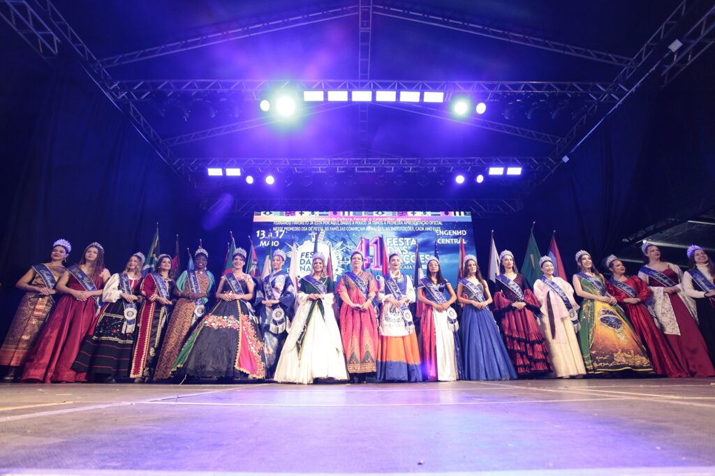 A imagem mostra um grupo de mulheres em um palco, alinhadas lado a lado, participando de um evento cultural ou concurso de beleza. Elas estão vestidas com roupas longas e elegantes, cada uma com uma faixa que indica o nome de diferentes países, como Tailândia, Portugal, México, Suíça, Brasil e Argentina, e usam coroas ou tiaras na cabeça. Ao fundo, há uma grande faixa que anuncia a "41ª Festa das Nações" no Engenho Central, em Piracicaba, SP, com datas de 13 a 17 de maio. O palco está iluminado com luzes azuis e brancas, criando um ambiente festivo e formal. Atrás das participantes, há diversas bandeiras nacionais, reforçando o tema multicultural do evento. A cena transmite uma celebração das culturas internacionais, com destaque para as representantes de diferentes nações.