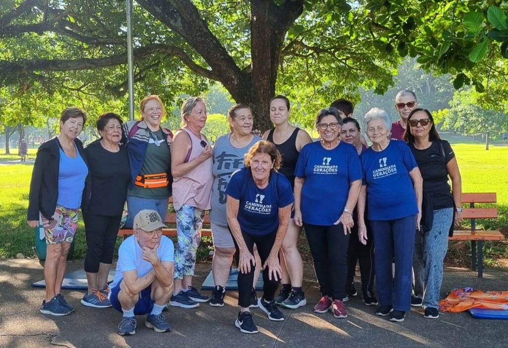A imagem mostra um grupo de 13 pessoas, predominantemente mulheres de meia-idade e idosas, reunidas ao ar livre em um parque ensolarado. O grupo está posicionado em frente a uma grande árvore, com um banco de madeira visível ao fundo. * A maioria das pessoas está vestida com roupas esportivas ou casuais, adequadas para uma atividade física. * Duas das participantes vestem camisetas azuis idênticas com a inscrição "CAMINHADA GERAÇÕES", sugerindo que fazem parte de um evento ou grupo organizado. * No centro, uma mulher está agachada com as mãos nos joelhos, enquanto um homem à esquerda está agachado na frente do grupo, com a mão no queixo. * O ambiente é um espaço público aberto, com gramado verde, luz natural intensa e sombras projetadas pelas árvores, indicando um dia claro. O clima parece ser de descontração e convívio social, típico de grupos de caminhada ou atividades físicas comunitárias para a terceira idade.