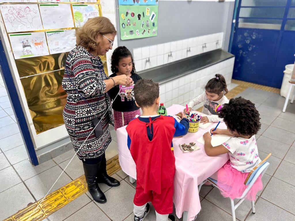 A imagem mostra uma cena em uma sala de aula ou ambiente escolar, onde uma professora está interagindo com quatro crianças ao redor de uma mesa coberta por uma toalha rosa. * A professora, uma mulher de cabelos castanhos claros e óculos, está de pé à esquerda, segurando um fio branco que se estende pelo ambiente. Ela veste um vestido estampado e botas pretas. * À direita, três crianças estão sentadas ou em pé ao redor da mesa desenhando ou colorindo. Uma menina de cabelos cacheados, usando uma blusa com estampas de borboletas e uma saia de tule rosa, está concentrada em seu papel. Ao lado dela, outra menina de camiseta branca observa a atividade. * De costas para a câmera, um menino veste uma fantasia de super-herói com capa vermelha e azul. Uma quarta criança, uma menina de camiseta preta, está de pé ao lado da professora, observando o que ela faz com o fio. * Sobre a mesa, há um pote com canetinhas coloridas, papéis e alguns pedaços de chocolate. Ao fundo, vê-se uma parede com desenhos infantis fixados e uma bancada com pias, típica de escolas. O ambiente é iluminado e organizado.