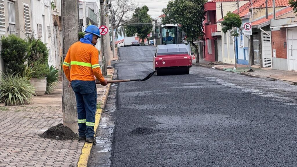A imagem mostra uma cena de trabalho na pavimentação de uma rua residencial. No primeiro plano, há um operário de costas, vestindo uma camiseta laranja com faixas refletoras amarelas, calça jeans e capacete azul, segurando uma pá. Ele está próximo a um poste de luz, em uma calçada de concreto. À frente, a rua está sendo recapeada com asfalto novo e escuro, e um rolo compactador vermelho está em operação, com um operador dentro da cabine. Ao redor, há casas de tijolos aparentes e algumas com pintura clara, típicas de um bairro tranquilo. Também é possível ver placas de trânsito, incluindo uma placa de “proibido estacionar”. O céu está nublado, indicando um dia cinzento. A imagem destaca o trabalho de infraestrutura urbana em andamento.