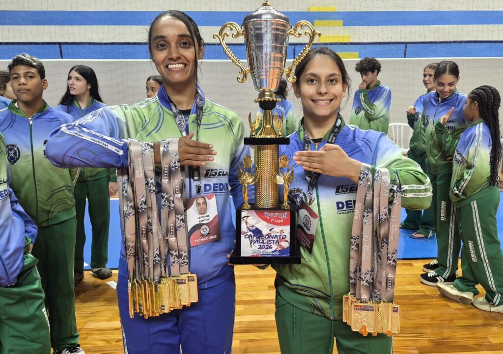 A imagem mostra uma cena de comemoração em um campeonato de taekwondo. No centro, duas mulheres vestindo uniformes esportivos nas cores verde e azul, com o logo "DEDINI", estão segurando juntas um grande troféu dourado. Ambas usam várias medalhas de ouro penduradas no pescoço, indicando várias conquistas. Uma delas tem um crachá que identifica seu nome como Leidiane Santos e a função de técnico para o ano de 2026. O troféu que seguram tem uma placa com a inscrição "Kyorugi 3º Lugar" e "Campeonato Paulista 2026 Taekwondo", mostrando que conquistaram o terceiro lugar na competição. Ao fundo, outros membros da equipe, também uniformizados, observam a cena, dentro de uma quadra esportiva coberta, com arquibancadas azuis e brancas. A atmosfera é de orgulho e celebração pelo desempenho no campeonato.