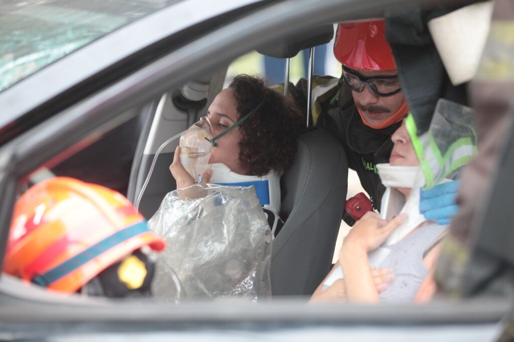 A imagem mostra uma cena de resgate ou atendimento de emergência dentro de um carro. Duas mulheres estão sentadas nos bancos dianteiros do veículo, ambas usando colares cervicais para imobilizar o pescoço, indicando que podem ter sofrido algum trauma. A mulher no banco do motorista está com uma máscara de oxigênio no rosto, segurando-a com a mão, e coberta por um plástico transparente, provavelmente para proteção ou isolamento. Um bombeiro, identificado pelo capacete vermelho e óculos de proteção, está inclinado para dentro do carro, aparentemente prestando auxílio ou avaliando o estado das vítimas. Outro capacete de bombeiro é visível no canto esquerdo da imagem, sugerindo a presença de mais profissionais no local. A cena transmite uma sensação de urgência e cuidado no atendimento às vítimas de um possível acidente.