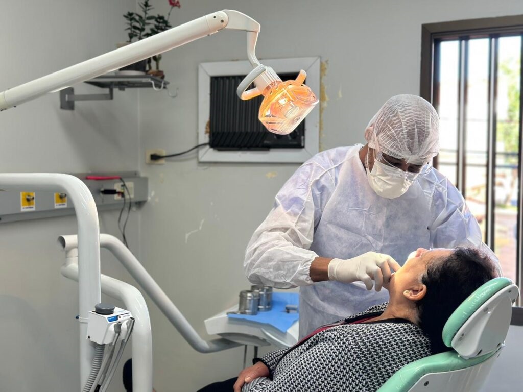 A imagem mostra uma cena em um consultório odontológico, onde um dentista realiza um atendimento clínico. * O profissional está posicionado à esquerda, utilizando equipamentos de proteção individual (EPIs) completos, incluindo gorro, máscara cirúrgica, óculos de proteção, luvas e avental descartável. Ele está inclinado sobre a paciente, realizando um procedimento na cavidade oral. * A paciente, uma mulher de cabelos escuros, está reclinada em uma cadeira odontológica de cor clara, mantendo uma postura de repouso enquanto recebe o tratamento. * O ambiente é iluminado por um foco de luz artificial direcionado para a boca da paciente. Ao fundo, observa-se uma parede com tomadas, uma janela com luz natural e uma prateleira com uma planta decorativa, caracterizando um espaço de atendimento médico funcional e organizado.
