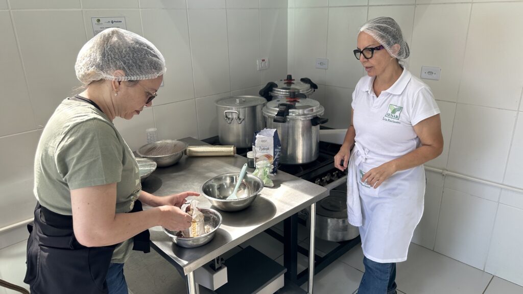 A imagem mostra uma cena em uma cozinha profissional ou institucional. Duas mulheres estão presentes, ambas usando toucas, o que indica um ambiente que segue normas de segurança alimentar. À esquerda, uma mulher vestindo uma camiseta verde-oliva está concentrada em um recipiente metálico, segurando um pacote pequeno enquanto prepara ingredientes. À direita, outra mulher, vestindo uma camisa polo branca com o logo do SENAR e um avental branco, observa a primeira mulher, possivelmente dando instruções ou colaborando. A cozinha possui superfícies de aço inoxidável, grandes panelas de pressão industriais sobre o fogão, e paredes revestidas de azulejos. No balcão central, há um pacote de farinha da marca Venturelli. A iluminação é clara e uniforme, típica de um ambiente interno bem iluminado. A perspectiva da foto está ao nível dos olhos, capturando a interação entre as duas pessoas no contexto de trabalho.