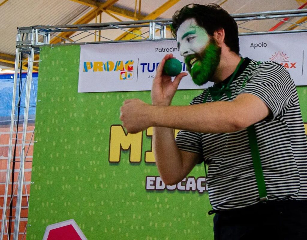 A imagem mostra um palhaço em uma apresentação, segurando uma bola verde próxima ao rosto com a mão esquerda. Ele veste uma camiseta listrada em preto e branco, suspensórios verdes e calças escuras. Seu rosto está pintado com maquiagem branca, com detalhes verdes ao redor dos olhos e uma barba e bigode verdes, combinando com o tema da roupa. O palhaço está sorrindo, com uma expressão animada e olhos focados na bola que segura. Ao fundo, há um grande banner verde com várias inscrições coloridas. Destacam-se as palavras "PROAC TU", "A FOI", "MAX M" e "ED.CAÇ", além das palavras "Patrocínio" e "Apoio" em uma faixa branca na parte superior do banner. O cenário sugere um evento cultural ou educacional, possivelmente relacionado a projetos apoiados pelo PROAC (Programa de Ação Cultural). A iluminação é clara e a imagem captura o movimento do palhaço, evidenciado pelo leve desfoque nas mãos.