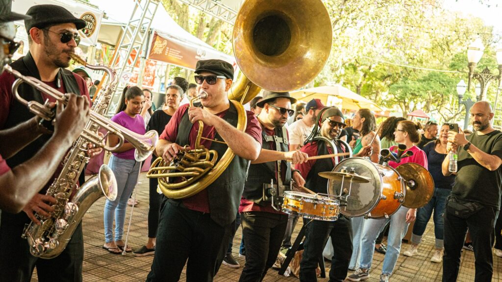 A imagem mostra uma banda de música de rua se apresentando ao ar livre, em um ambiente que parece ser uma praça ou evento público. * No centro, um músico toca um sousafone (tuba grande), enquanto à esquerda outro toca um saxofone. À direita, um percussionista toca uma bateria adaptada, composta por uma caixa e um bumbo, acompanhado por um músico que toca um prato. * Os músicos estão vestidos de forma semelhante, usando camisas bordô, coletes pretos, chapéus e óculos escuros, conferindo um estilo uniforme ao grupo. * Ao fundo, várias pessoas assistem à apresentação. Algumas estão paradas observando, enquanto outras caminham ou conversam. Um homem à direita segura um celular, sugerindo que está gravando ou fotografando a performance. * O ambiente é iluminado pela luz do dia, com árvores e postes de iluminação visíveis ao fundo, criando uma atmosfera descontraída e festiva.