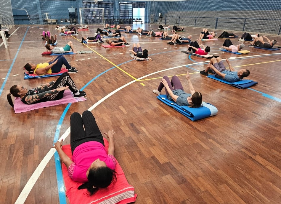 A imagem mostra uma aula de ginástica em grupo realizada dentro de um ginásio esportivo. Várias pessoas, principalmente adultos, estão deitados de costas em colchonetes distribuídos pelo chão de madeira da quadra. Elas parecem estar realizando exercícios de fortalecimento abdominal, com os troncos elevados e braços esticados para frente. Os participantes vestem roupas esportivas coloridas, como rosa, azul, preto e amarelo. O ambiente é amplo, com teto alto, paredes com redes e uma trave de futebol ao fundo, indicando que o local é um ginásio multifuncional. A iluminação é clara, e a foto foi tirada de um ângulo elevado, mostrando a organização dos alunos no espaço. A atmosfera é de concentração e atividade física em grupo.
