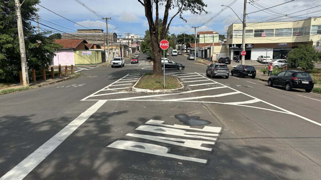 A imagem mostra um cruzamento urbano durante o dia, com uma ilha central que possui uma árvore grande e um placa de trânsito com a palavra "PARE". No chão, também está pintado "PARE" em letras grandes, indicando a necessidade de parada dos veículos. Há vários carros na rua, incluindo um caminhonete branca, um sedã escuro, um hatch prata e outro veículo preto. Ao fundo, há prédios comerciais, com placas visíveis, como "LS" e "Lumina". Também é possível ver uma placa de venda com um número de telefone. O céu está parcialmente nublado, e a iluminação natural cria sombras das árvores e dos prédios sobre o asfalto. A cena parece ser em um bairro movimentado, com trânsito moderado e pedestres circulando.