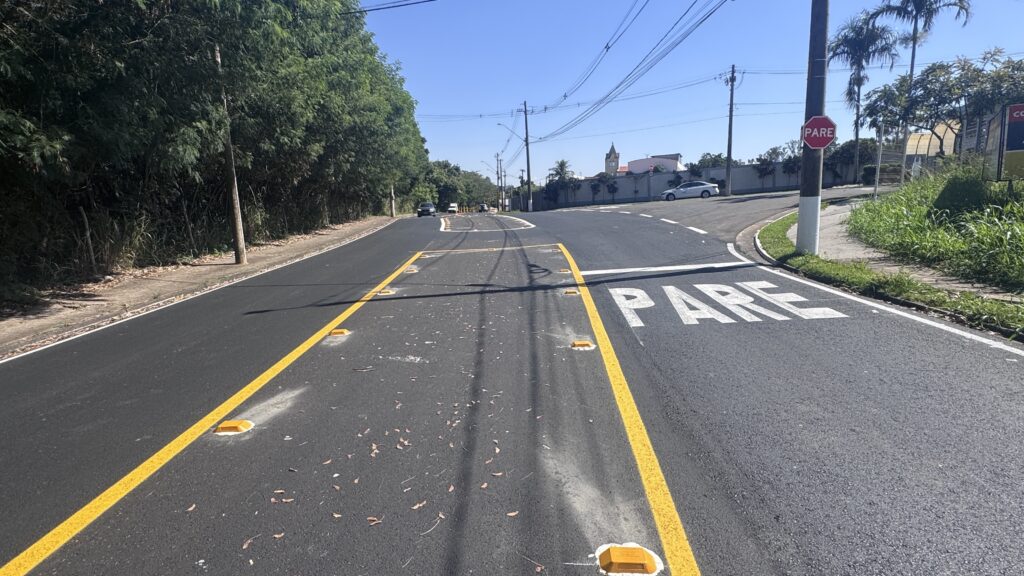 A imagem mostra uma estrada asfaltada em um dia ensolarado. No centro da imagem, há uma faixa de pedestres pintada no chão com a palavra "PARE" em letras grandes e brancas, indicando que os veículos devem parar. À direita, há uma calçada com grama verde e um poste com uma placa de pare vermelha, reforçando a sinalização de parada. A estrada segue em direção a um cruzamento, onde é possível ver algumas árvores e um carro branco ao fundo. O céu está claro e azul, e a iluminação forte sugere que é durante o dia. A imagem é capturada de um ângulo baixo, próximo ao chão, destacando as marcações da pista e a sinalização de trânsito.