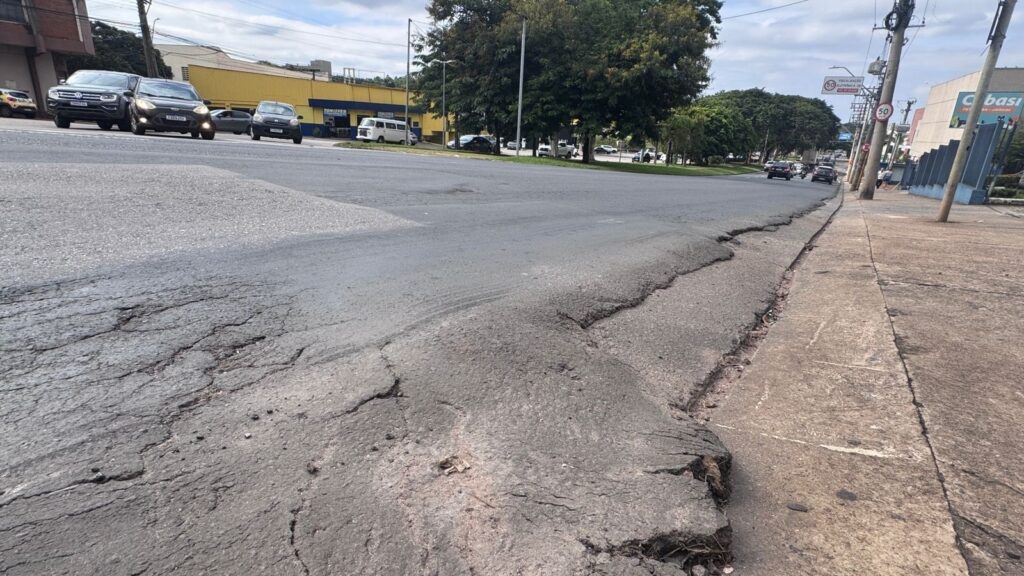 A imagem mostra uma rua em uma área urbana, vista de um ângulo baixo, com grande foco no pavimento danificado em primeiro plano. O asfalto está muito deteriorado, com rachaduras profundas, buracos e partes desfeitas, criando um desnível acentuado em relação à calçada de concreto ao lado. Ao fundo, é possível ver uma avenida com várias faixas, por onde circulam alguns veículos, incluindo um SUV escuro, um carro pequeno e uma van branca. Há um poste na calçada à direita, e um prédio comercial com uma placa parcialmente visível, onde se lê "Cbasi". Também há placas de sinalização indicando fiscalização eletrônica de velocidade com limite de 50 km/h. O céu está nublado, sugerindo um dia cinzento. A imagem destaca principalmente o estado precário da pavimentação da via.