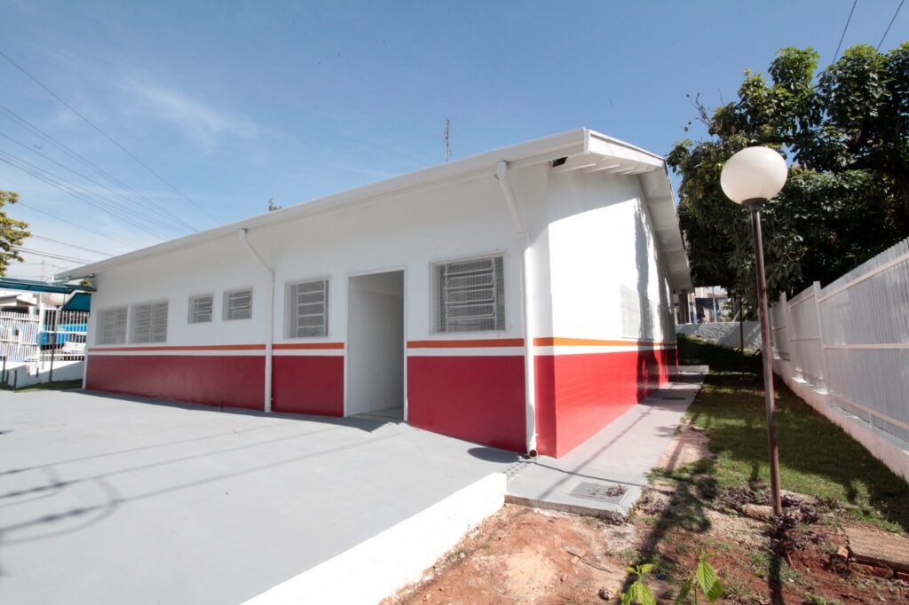 A imagem mostra um prédio de um andar, com um design simples e moderno, provavelmente uma escola ou algum tipo de instalação comunitária. A fachada do prédio é pintada em branco com uma faixa horizontal laranja e uma parte inferior vermelha. Há várias janelas com grades de metal brancas e uma porta aberta ao centro. O telhado é branco e se estende além das paredes, criando uma pequena área de sombra na frente. O chão em frente ao prédio é de concreto liso. À direita, pode-se ver um poste de luz redondo, um pequeno gramado e uma cerca branca. O céu está claro e azul, sugerindo um dia ensolarado e sem nuvens. A imagem transmite uma sensação de limpeza, organização e simplicidade.