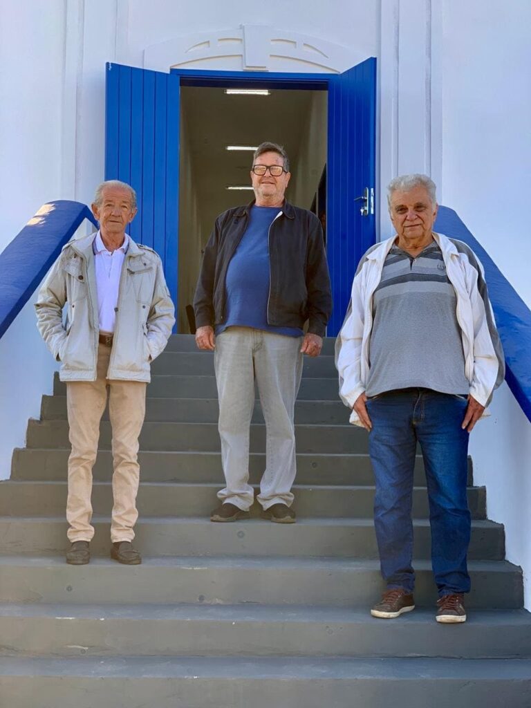 A imagem mostra três homens adultos em pé na escadaria frontal de um edifício com arquitetura estilo colonial ou mediterrânea, caracterizado por paredes brancas e uma porta de entrada dupla em um tom vibrante de azul. * O homem à esquerda veste uma jaqueta clara, camisa pólo branca e calças bege, com uma postura ereta e as mãos nos bolsos. * O homem ao centro é mais alto, usa óculos de grau, uma jaqueta escura sobre uma camiseta azul e calças cinzas, posicionando-se um degrau abaixo dos outros. * O homem à direita veste uma jaqueta branca sobre uma camisa polo cinza listrada e calças jeans azuis, mantendo uma postura relaxada. A iluminação é natural e abundante, sugerindo um dia ensolarado, o que destaca as cores vivas da fachada e cria sombras suaves sob os degraus cinzas.