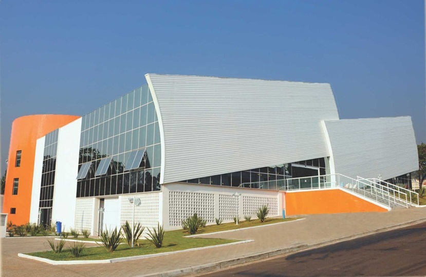 A imagem mostra um edifício moderno de múltiplos andares com um design arquitetônico marcante. A fachada combina elementos curvos e angulares, com grandes painéis de vidro que permitem ver o interior do prédio. O teto e as paredes superiores são feitos de metal ondulado, em tom cinza metálico. Um destaque visual é uma seção cilíndrica pintada de laranja vibrante no lado esquerdo do edifício, que contrasta com as paredes brancas e o metal cinza. Na parte inferior, há uma parede branca com um padrão vazado, que adiciona textura à construção. O céu azul claro ao fundo realça as cores do edifício, sugerindo que a foto foi tirada durante o dia, provavelmente no final da manhã ou início da tarde. A iluminação cria sombras suaves que valorizam os detalhes arquitetônicos. Na frente do prédio, há um gramado verde com plantas espinhosas e um caminho pavimentado que leva até a entrada, acessível por uma rampa e uma escada laranja. Também é possível ver uma lixeira azul próxima à calçada. O ambiente parece ser urbano ou suburbano, possivelmente um complexo comercial ou institucional, e não há pessoas visíveis na cena. A perspectiva da foto é ligeiramente elevada, mostrando o prédio de frente e de lado, com uma composição equilibrada que destaca as formas e materiais diferentes da construção.
