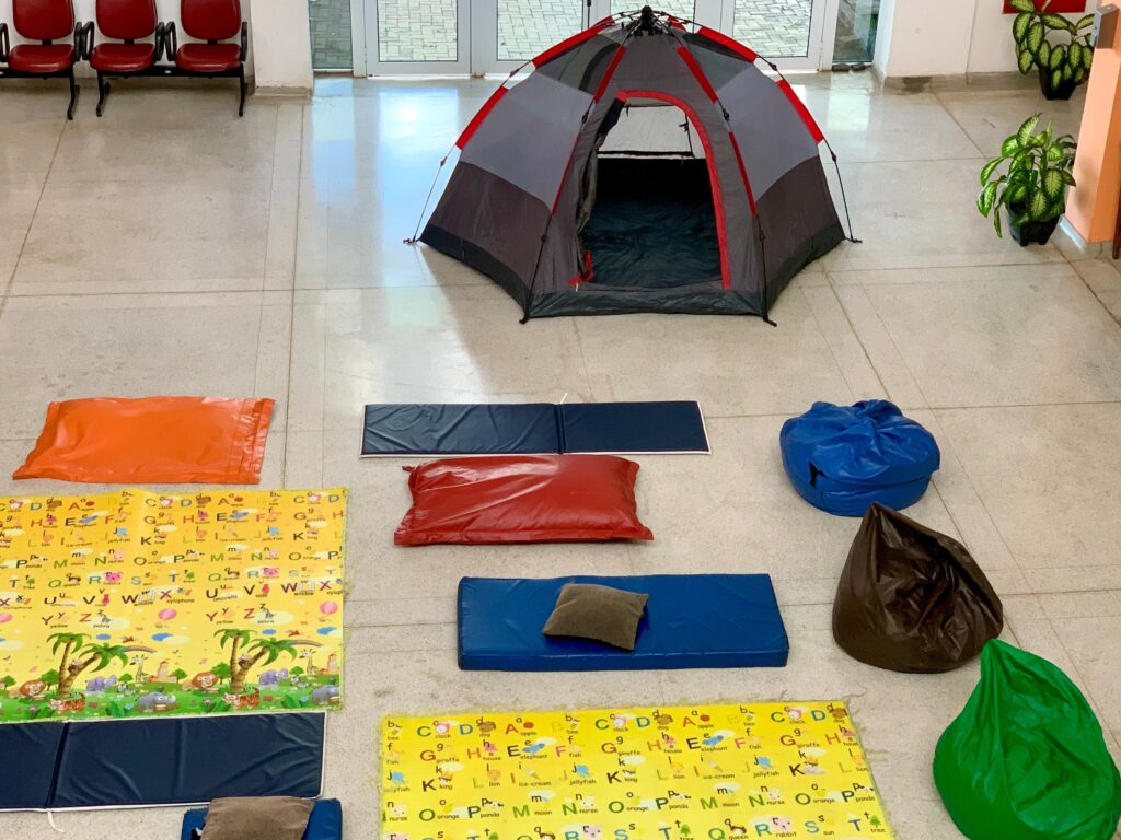 A imagem mostra uma área interna, possivelmente uma sala de recreação infantil ou um espaço de atividades, organizada com vários itens de conforto e lazer. * No centro superior, há uma barraca de camping montada, com estrutura cinza e detalhes em vermelho. * O piso está coberto por diversos tapetes educativos amarelos, estampados com o alfabeto e ilustrações de animais, além de vários colchonetes retangulares nas cores azul e laranja. * Espalhados pelo espaço, encontram-se vários pufes de diferentes cores, incluindo azul, marrom e verde, além de almofadas pequenas sobre os colchonetes. * No canto superior esquerdo, há fileiras de cadeiras vermelhas, e no lado direito, algumas plantas em vasos decoram o ambiente, que possui um piso de cerâmica clara e portas de vidro ao fundo.