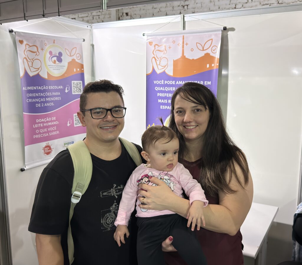 Essa foto é muito bonita e registra um momento especial! Nela, vemos uma família simpática — pai, mãe e um bebê — posando em um evento ou feira. O pai, que está à esquerda, usa óculos e uma camiseta de moto, enquanto a mãe segura o bebê, que está uma fofura com uma blusa da Minnie Mouse. Eles estão em frente a alguns banners educativos sobre aleitamento materno e saúde infantil, o que sugere que eles estão em um ambiente informativo, talvez em uma campanha ou evento comunitário