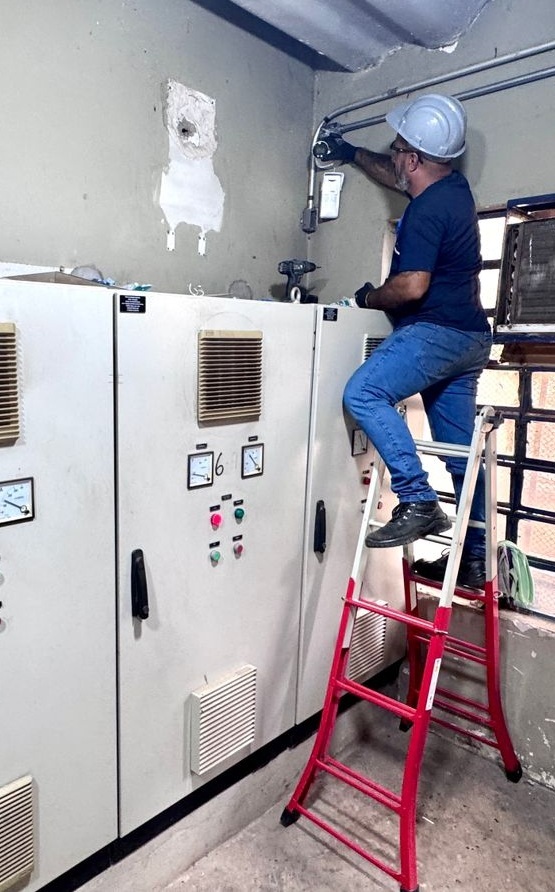 Uma fotografia vertical que mostra um técnico a realizar manutenção/instalação elétrica num ambiente industrial/comercial. Elementos principais Técnico: usa capacete branco, t-shirt escura e calças de ganga; está em cima de um escadote vermelho a trabalhar junto a condutas/eletrodutos. Escadote: robusto, vermelho, com degraus antiderrapantes. Painel elétrico: armários metálicos bege com grelhas de ventilação, mostradores analógicos (voltímetro/amperímetro) e botões de comando (verdes e vermelhos). Um botão tem o número “6” marcado. Ferramentas: uma parafusadora/berbequim sem fios pousada sobre o painel. Ambiente: parede com sinal de intervenção (mancha de cimento/gesso) e janela gradeada à direita. Observações relevantes Formato e qualidade: fotografia digital vertical, de boa resolução e nítida; detalhes visíveis. Legibilidade: imagem clara — não há texto extenso para ler (apenas o “6” marcado). Segurança: mostra trabalho em altura; a imagem sugere que há procedimentos de segurança em curso (capacete, escadote), mas não é possível confirmar cumprimento total das normas só pela foto.