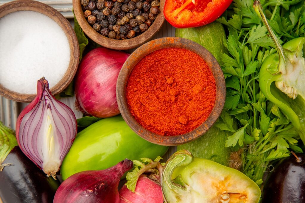 A imagem mostra uma vista aérea de uma mesa de madeira clara onde estão dispostos vários ingredientes frescos e temperos, organizados de forma harmoniosa. No centro, há uma tigela de madeira cheia de pó vermelho, que parece ser páprica ou pimenta em pó. Ao redor, há outras tigelas menores: uma com sal grosso e outra com grãos de pimenta-do-reino inteiros. Também estão presentes vegetais frescos, como cebolas roxas (uma inteira e uma cortada ao meio), um rabanete, um pimentão verde inteiro e outro cortado ao meio, uma abobrinha e um ramo de salsa fresca. A iluminação é clara e destaca as cores vibrantes e as texturas naturais dos ingredientes, criando uma composição visualmente atraente e convidativa, ideal para um ambiente culinário.