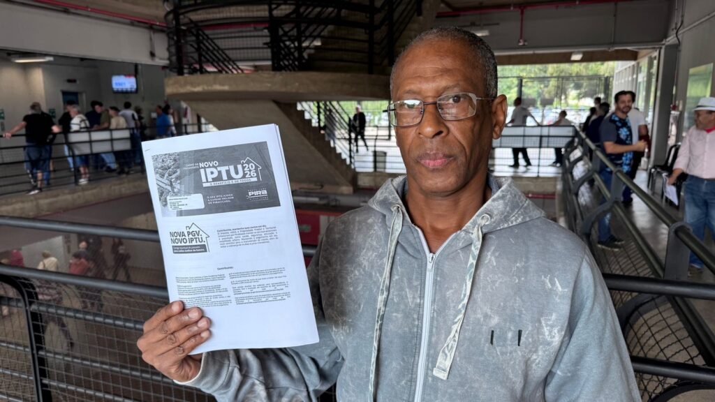 A imagem mostra um homem de meia-idade, de pele negra e óculos, posicionado em primeiro plano, olhando diretamente para a câmera. Ele veste um moletom cinza com zíper e segura, com a mão esquerda, um documento oficial da Prefeitura de Piracicaba, identificado como o carnê de pagamento do IPTU 2026. O documento, que ocupa parte significativa da imagem, apresenta o logotipo da prefeitura e frases como "O benefício é de todos" e "Nova PGV. Novo IPTU". O cenário de fundo sugere ser o interior de um prédio público ou rodoviária, com uma estrutura metálica, escadas e corrimãos. É possível observar outras pessoas transitando ao fundo, tanto no nível inferior quanto no superior, criando um ambiente movimentado e cotidiano. A iluminação é natural, proveniente de grandes aberturas na parte superior da estrutura.