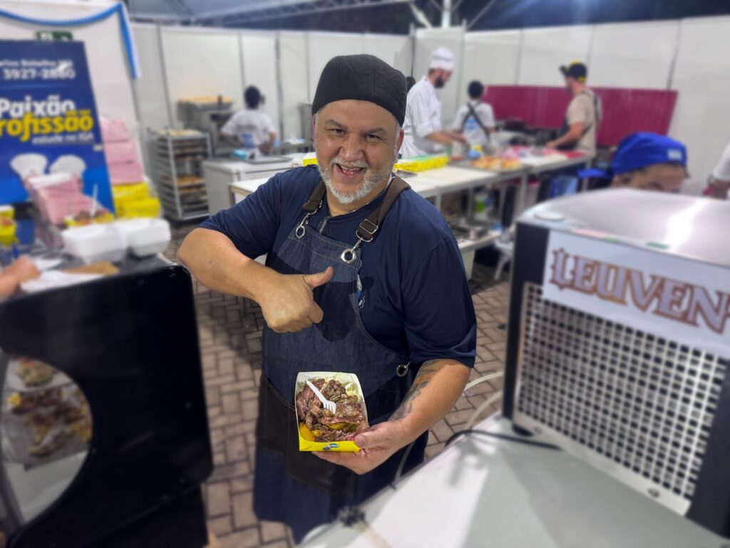 A imagem mostra um homem sorridente, provavelmente um chef, que está dando um sinal de positivo com o polegar da mão direita. Ele veste uma camiseta azul escura, avental marrom e um gorro preto. Ele tem barba grisalha e uma tatuagem visível no antebraço esquerdo. Na mão esquerda, ele segura uma pequena bandeja amarela com pedaços de carne grelhada fatiada e um garfo branco. O cenário parece ser uma cozinha industrial ou uma barraca de comida ao ar livre, com outras pessoas trabalhando ao fundo, também vestidas com aventais e gorros. O ambiente é iluminado artificialmente, sugerindo que a foto foi tirada à noite. No canto esquerdo da imagem, há uma placa azul com o texto "Paixão profissão estude no IGA LEUVEN" e, no canto direito, uma parte de um equipamento com a palavra "LEUVEN". A cena transmite uma atmosfera de profissionalismo e entusiasmo pela culinária, destacando a ligação entre paixão e profissão.