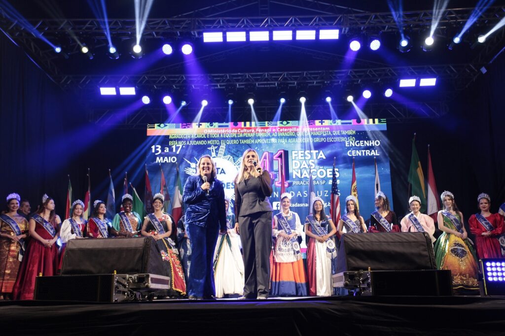 A imagem mostra um evento cultural ou concurso de beleza chamado "41ª Festa das Nações", realizado em Piracicaba, SP, em maio de 2016. No palco, duas mulheres estão no centro, ambas vestidas de forma elegante e segurando microfones, possivelmente apresentadoras ou anfitriãs do evento. Atrás delas, há uma fila de jovens mulheres usando coroas e faixas, representando diferentes nações, e vestindo trajes típicos ou roupas formais que refletem suas culturas. O palco é grande e bem iluminado, com luzes azuis e holofotes que destacam os participantes. Ao fundo, há uma grande tela digital que exibe o nome do evento, as datas (13 a 17 de maio), e agradecimentos a patrocinadores e colaboradores, como o Ministério da Cultura, Fenapi, Caterpillar, entre outros. Também é possível ver várias bandeiras nacionais dispostas em um suporte ao fundo, reforçando o tema multicultural da festa. A atmosfera geral é de celebração e formalidade, com um público provavelmente presente além da área visível na foto. O evento parece ser uma festa tradicional que homenageia a diversidade cultural através da apresentação de representantes de diferentes países.