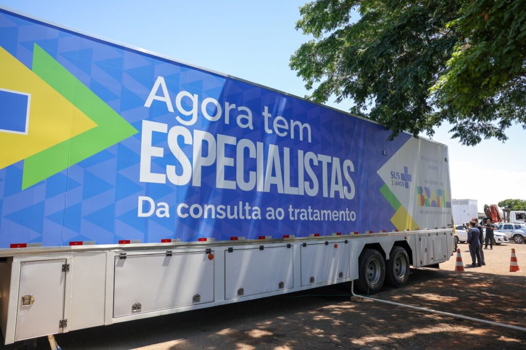 A imagem mostra um caminhão-baú branco estacionado em uma área aberta, possivelmente um terreno de terra batida. Na lateral do caminhão há uma grande faixa azul com detalhes em verde e amarelo, cores da bandeira do Brasil, onde está escrito em letras brancas: "Agora tem ESPECIALISTAS" e "Da consulta ao tratamento". Também aparecem os logos do Ministério da Saúde, do SUS (Sistema Único de Saúde) com a marca dos 35 anos, e do Governo do Brasil, acompanhados do slogan "Do lado do povo brasileiro". Ao lado do caminhão, há algumas pessoas em pé, incluindo um homem de terno, e ao fundo há um carro branco estacionado. O ambiente é ao ar livre, com árvores ao redor e luz do sol forte, indicando que a foto foi tirada durante o dia. A imagem sugere uma ação do governo relacionada à saúde, possivelmente um caminhão móvel para atendimento médico especializado.