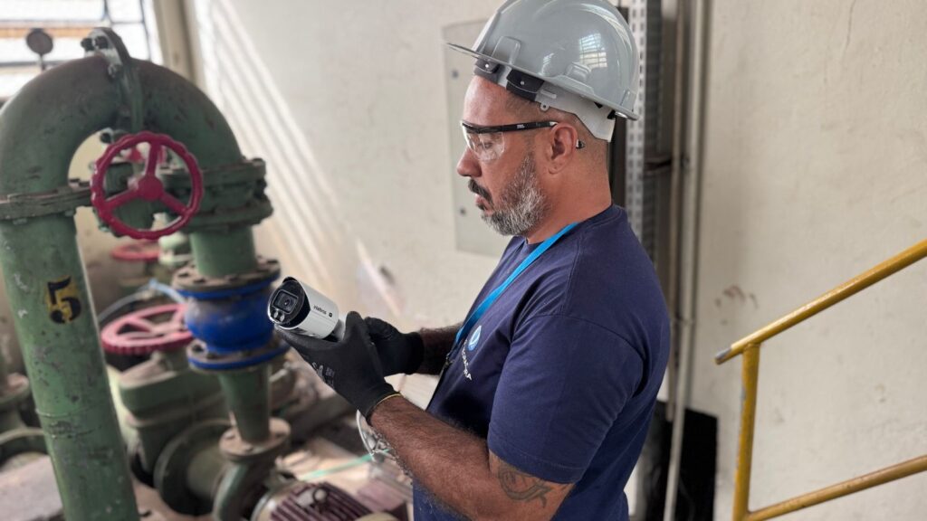 A imagem mostra um técnico, possivelmente um engenheiro ou instalador, em um ambiente industrial. Ele está de perfil, usando capacete branco, óculos de proteção transparentes, luvas pretas e uma camiseta azul com um logo. O homem está segurando uma câmera de segurança do tipo bullet, da marca Intelbras, que parece estar sendo instalada ou ajustada. Ao fundo, há uma tubulação grande pintada de verde, com uma válvula vermelha e o número 5 pintado em amarelo. A tubulação parece ser parte de uma instalação industrial, como uma fábrica ou uma planta de tratamento. Também é possível ver uma grade amarela e uma parede clara ao fundo, indicando que o local é interno ou parcialmente coberto. A imagem transmite uma sensação de trabalho técnico e cuidado com a segurança, destacando o profissionalismo do técnico e o ambiente industrial onde a tecnologia de segurança está sendo aplicada.