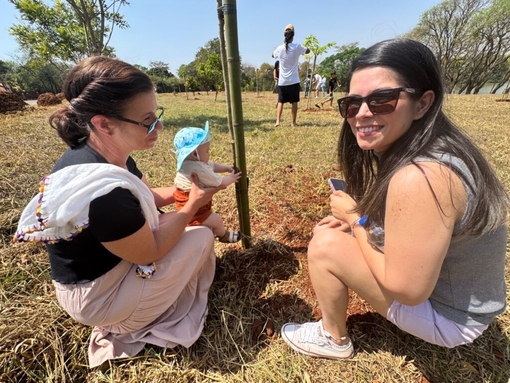 A imagem mostra uma cena ao ar livre, provavelmente durante um evento comunitário de plantio de árvores, em um dia ensolarado. Em primeiro plano, duas mulheres estão agachadas sobre um terreno seco e com grama rala. A mulher à esquerda, vestindo uma camiseta preta e uma saia clara, segura um bebê que usa um chapéu azul e shorts laranja; o bebê está alcançando um pequeno bambu que sustenta uma muda recém-plantada. A mulher à direita está sorrindo para a câmera, usando óculos escuros, regata cinza e shorts claros, segurando um celular. Ao fundo, outras pessoas estão espalhadas pelo campo, também participando do plantio de árvores. O céu está claro e azul, e a luz do sol é forte, criando sombras no chão. Parece ser um parque ou uma área aberta com poucas árvores ao redor. A imagem transmite uma sensação de colaboração, natureza e cuidado ambiental.