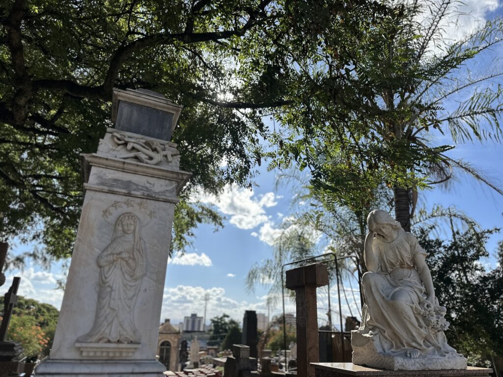 A imagem mostra um cemitério tranquilo sob um céu azul com algumas nuvens. No primeiro plano, há duas esculturas de mármore impressionantes. À esquerda, uma estátua alta e ornamentada retrata uma figura vestida com uma túnica, possivelmente um santo, com um halo de estrelas ao redor da cabeça e um desenho de âncora na parte superior do monumento. À direita, uma escultura expressa tristeza: uma mulher sentada, com a cabeça abaixada e a mão na testa, segurando um buquê de flores, vestindo roupas fluidas que parecem estar caindo suavemente. Ao redor das esculturas, há árvores verdes que criam um jogo de luz e sombra no chão e nas estátuas, transmitindo uma atmosfera calma e serena. Ao fundo, é possível ver várias lápides e monumentos, indicando que o cemitério é antigo e extenso. Além do cemitério, na distância, aparecem edifícios que fazem parte de uma cidade. A perspectiva da imagem é de baixo para cima, dando uma sensação de grandiosidade e solenidade, reforçando o sentimento de respeito e memória que o local inspira. A cena geral é contemplativa, transmitindo paz e reflexão sobre a vida e a morte.