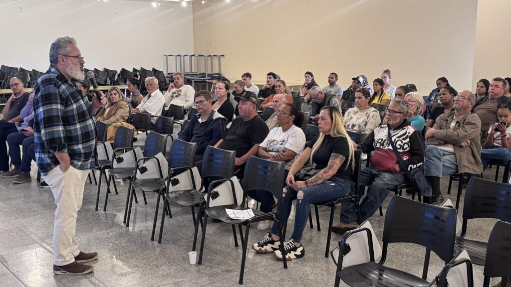A imagem mostra um encontro em ambiente interno, possivelmente uma palestra, workshop ou treinamento. Um homem de meia-idade, com barba grisalha e óculos, veste uma camisa xadrez azul e preta com calça clara, está em pé segurando um microfone e falando para o público. As pessoas sentadas, de diferentes idades, estão organizadas em cadeiras pretas de plástico, prestando atenção ao palestrante. O ambiente é simples, com paredes claras, piso de azulejo e iluminação embutida no teto. Ao fundo, há várias cadeiras empilhadas, sugerindo que o espaço é multiuso. A foto foi tirada de lado, mostrando tanto o palestrante quanto parte da plateia, destacando a interação entre eles.