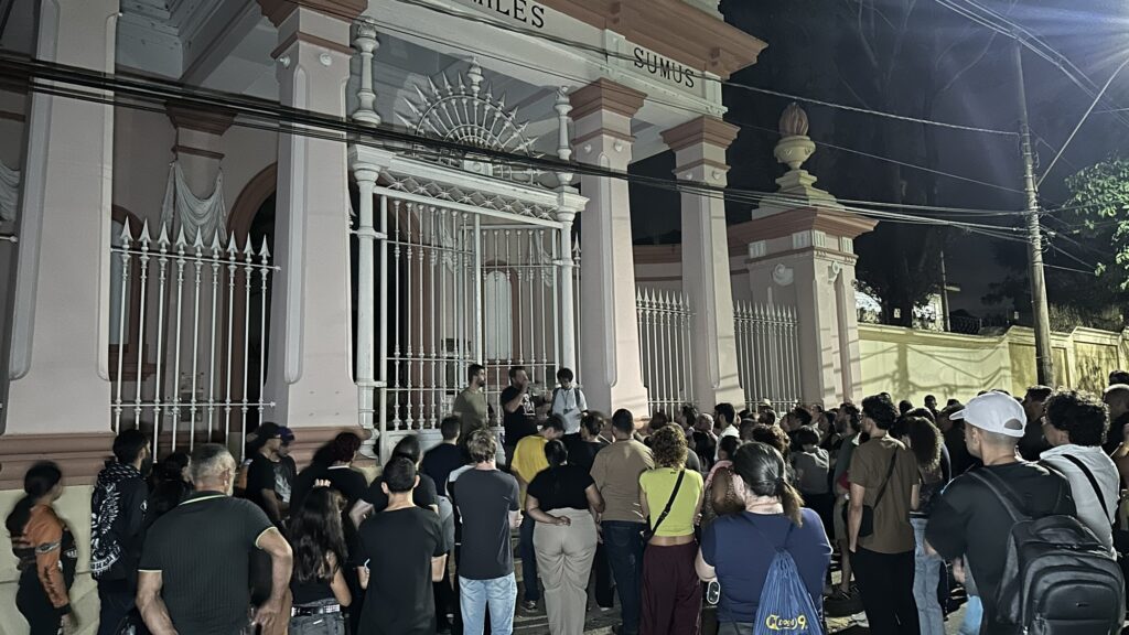 A imagem mostra uma cena noturna em frente a um portão monumental e histórico, provavelmente a entrada de um cemitério ou um prédio institucional antigo. A construção apresenta elementos arquitetônicos clássicos, como colunas altas e uma fachada em tons claros, com detalhes em rosa e branco. No topo do portão, há uma grade de ferro decorativa com um desenho que lembra raios de sol, e as palavras "LES" e "SUMUS" estão inscritas na parte superior da estrutura. Na frente do portão, há um grupo de pessoas reunidas, algumas parecem estar conversando ou ouvindo alguém, sugerindo que pode ser uma visita guiada ou um evento. A maioria está vestida de forma casual, com camisetas, calças jeans e mochilas. A iluminação é artificial, provavelmente de postes de luz, conferindo um tom quente e amarelo à cena. Fios elétricos cruzam a parte superior da imagem, destacando ainda mais o cenário noturno. A atmosfera geral é de concentração e interesse em relação ao local histórico ou cultural que está sendo visitado.