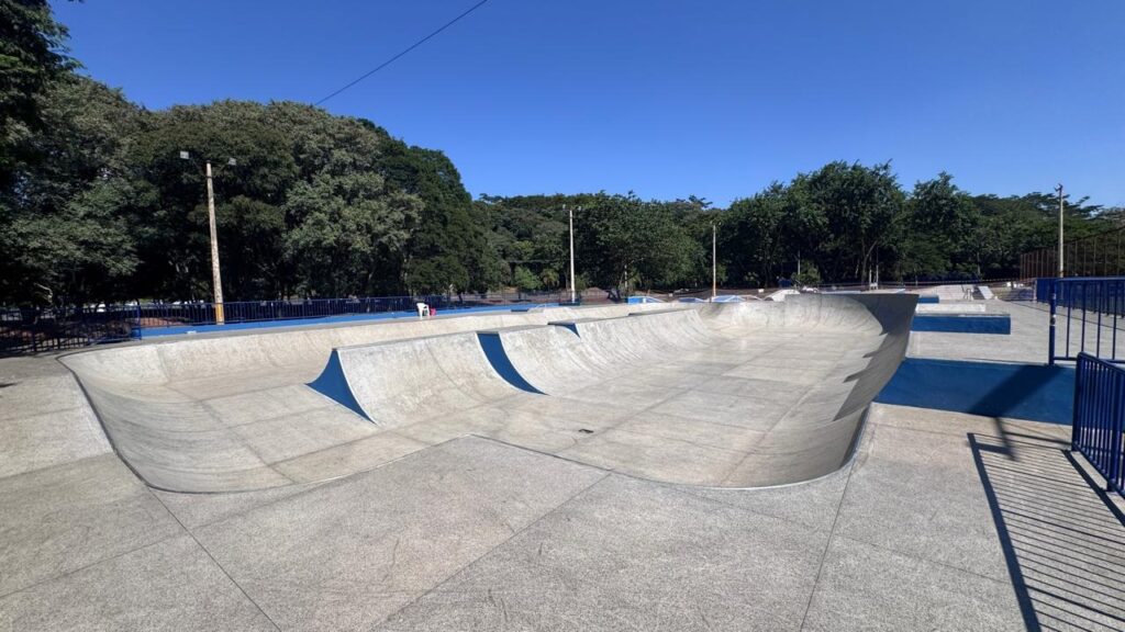 A imagem mostra um parque de skate amplo e vazio, feito de concreto, sob um céu azul claro e sem nuvens. O parque apresenta diversas rampas, bowls e bordas, todas feitas de concreto cinza claro, com detalhes em azul nas bordas e algumas superfícies planas. A iluminação é intensa, indicando um dia ensolarado, e as sombras projetadas pelas estruturas e por uma cerca próxima são bem definidas. O concreto tem uma textura áspera, típica de parques de skate, com linhas e divisões visíveis que delimitam as diferentes áreas. Ao fundo, há uma linha densa de árvores verdes, com algumas mais altas à esquerda e uma fileira mais uniforme à direita, sugerindo que o parque está situado em um ambiente natural ou de parque urbano. Também é possível ver alguns postes de iluminação entre as árvores. Não há pessoas na imagem, o que traz uma sensação de tranquilidade e expectativa para o uso do espaço.