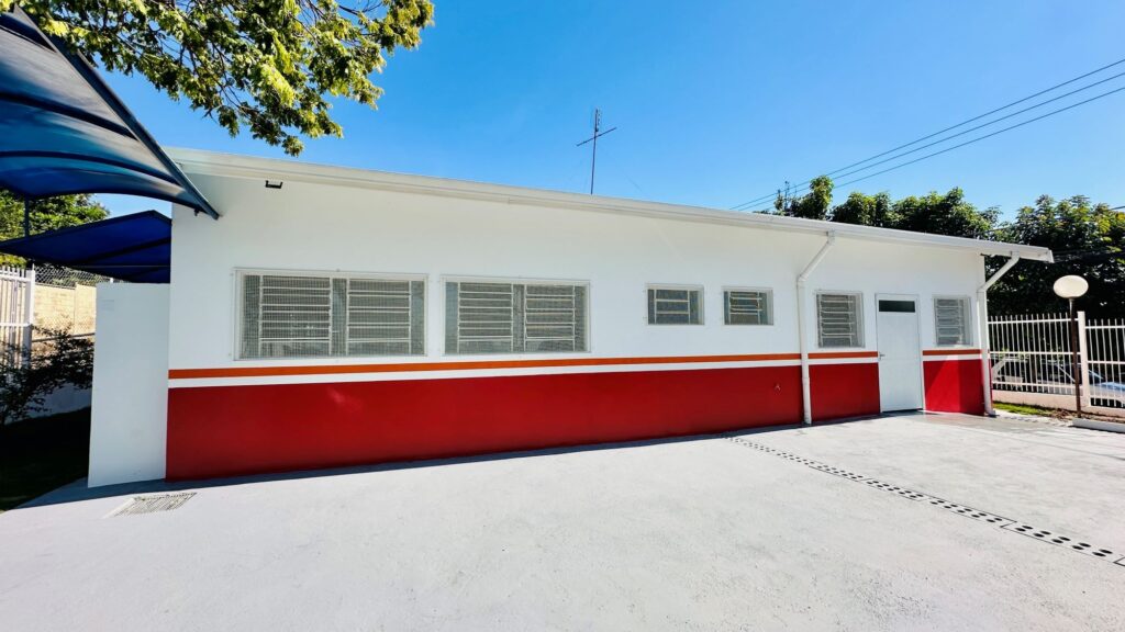 A imagem apresenta a fachada lateral de uma edificação térrea de arquitetura simples, pintada de branco com uma faixa horizontal vermelha e uma linha fina alaranjada na parte inferior das paredes. * A estrutura possui diversas janelas retangulares protegidas por grades metálicas brancas e uma porta de entrada centralizada. * O telhado é de uma água, com calhas brancas aparentes, e há uma cobertura externa azul à esquerda. * O piso é cimentado, de cor cinza claro, com um sistema de drenagem linear visível no chão. * O céu está limpo e azul, com parte de uma árvore à esquerda e uma cerca de metal branca delimitando o terreno ao fundo.