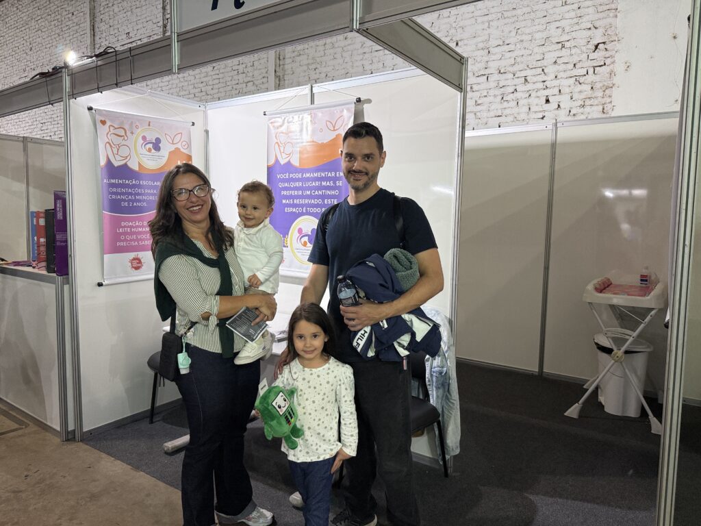 A imagem mostra uma família de quatro pessoas posando para uma foto em um ambiente interno, provavelmente em uma feira ou evento relacionado à saúde e amamentação. À esquerda, uma mulher segura uma criança pequena vestida de branco. No centro, uma menina segura um brinquedo verde, e à direita, um homem segura uma garrafa de água e uma jaqueta, com o braço apoiado na menina. Ao fundo, há dois banners com informações em português sobre alimentação escolar para crianças menores de 2 anos, doação de leite humano e incentivo à amamentação em qualquer lugar, além de um espaço reservado para amamentar. O ambiente é iluminado por luz artificial e tem paredes brancas. A atmosfera transmite uma sensação de cuidado, apoio e informação para famílias com crianças pequenas.