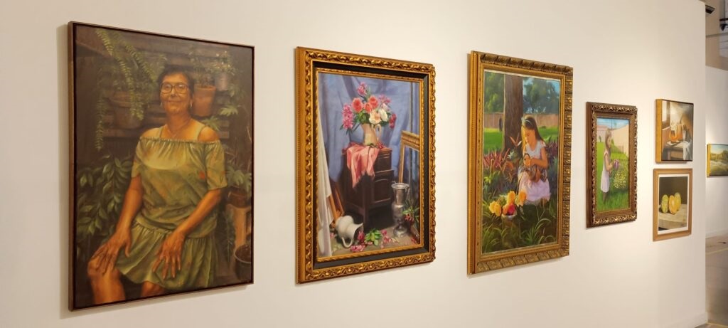 A imagem mostra uma exposição de pinturas em uma galeria de arte, com cinco obras penduradas em uma parede branca. Da esquerda para a direita: Um retrato grande de uma mulher idosa com óculos, vestindo um vestido verde esmeralda, sentada em um ambiente com muitas plantas, transmitindo uma sensação de tranquilidade e natureza. Uma pintura de natureza morta com um vaso de flores coloridas, um tecido branco e alguns objetos metálicos, tudo sobre uma superfície azul. Uma paisagem com uma menina jovem vestida de branco, sentada em um jardim ensolarado, cercada por árvores e flores. Um retrato menor de uma menina em um ambiente ao ar livre, possivelmente em um jardim. Dois quadros pequenos empilhados na extremidade direita, um mostrando frutas ou objetos sobre uma mesa e o outro uma imagem próxima de frutas cítricas. As molduras são variadas: as maiores são douradas e ornamentadas, enquanto as menores têm molduras mais simples em tons de madeira. A iluminação é clara e uniforme, destacando as cores e detalhes das pinturas. O ambiente transmite uma atmosfera calma e contemplativa, típica de uma galeria de arte.