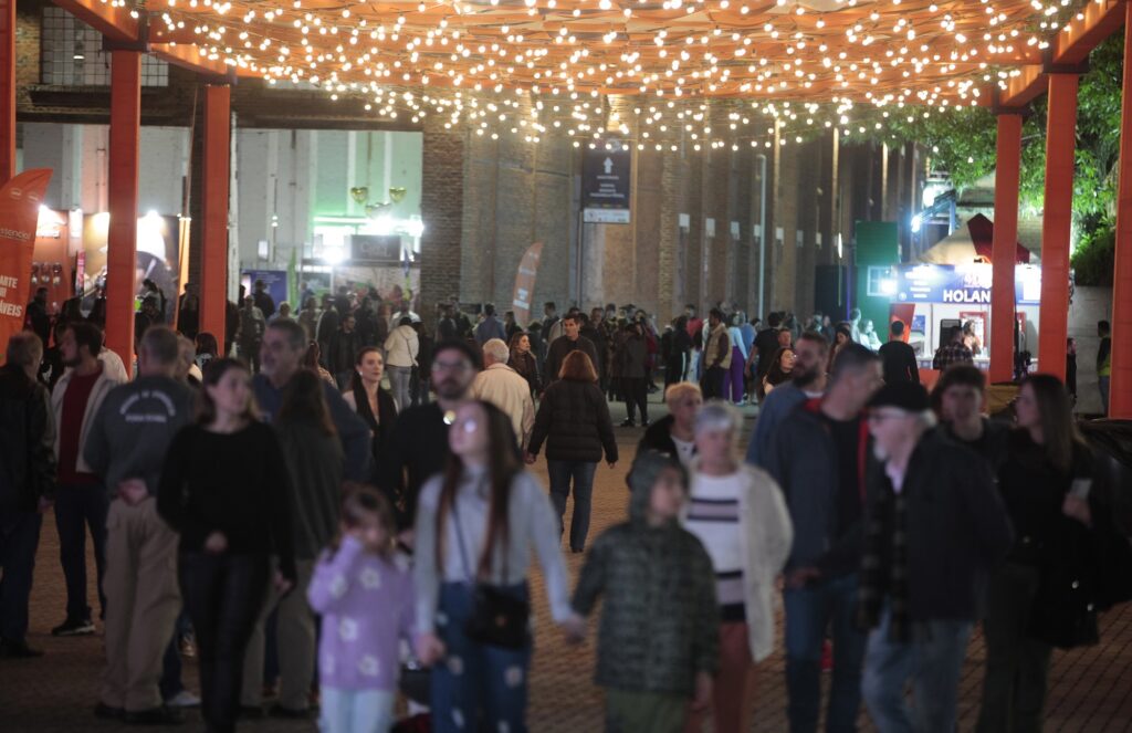 A imagem mostra uma rua ao ar livre à noite, cheia de pessoas caminhando em diferentes direções. A rua é decorada com muitas luzes de cordão penduradas no alto, criando uma iluminação quente e acolhedora que ilumina todo o ambiente com um tom dourado. As pessoas estão vestidas com roupas casuais, algumas com jaquetas e suéteres, indicando que o clima pode estar fresco. A rua é ladeada por grandes colunas laranjas e prédios de tijolos ao fundo. Há barracas ou estandes ao longo da rua, sugerindo que pode ser um mercado noturno ou uma festa de rua. O ambiente parece movimentado e animado, com um clima social e festivo. A palavra "HOLAN" aparece em uma placa ao fundo, possivelmente o nome de um estabelecimento ou do evento.