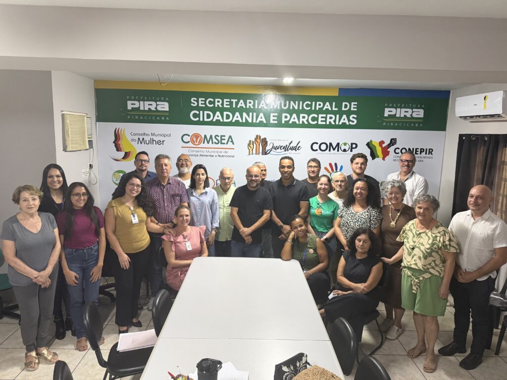 A imagem mostra um grupo de cerca de 25 pessoas reunidas em um ambiente interno, provavelmente um escritório ou sala de reuniões. Elas estão posicionadas ao redor e atrás de uma grande mesa branca. Ao fundo, há uma faixa ou banner grande fixado na parede com diversos logos e textos, entre eles "SECRETARIA MUNICIPAL DE CIDADANIA E PARCERIAS", "PREFEITURA PIRACICABA" e nomes de conselhos municipais, como o "Conselho Municipal da Mulher", "COMSEA", "COMOP" e "Conselho do Orçamento Participativo". A iluminação é clara e uniforme, provavelmente proveniente de luzes fluorescentes no teto. As pessoas estão sorrindo e parecem estar em um momento de confraternização ou celebração. Elas apresentam diversidade de idade e gênero, vestindo roupas casuais a semi-formais. Algumas estão sentadas à mesa, outras em pé, todas voltadas para a câmera. No canto superior direito da parede, há um aparelho de ar-condicionado. A foto foi tirada de uma posição um pouco mais baixa, com uma leve perspectiva para cima. O ambiente transmite a sensação de um encontro oficial ou reunião de um grupo ligado à administração pública municipal de Piracicaba, relacionado a temas de cidadania, parcerias e conselhos comunitários.