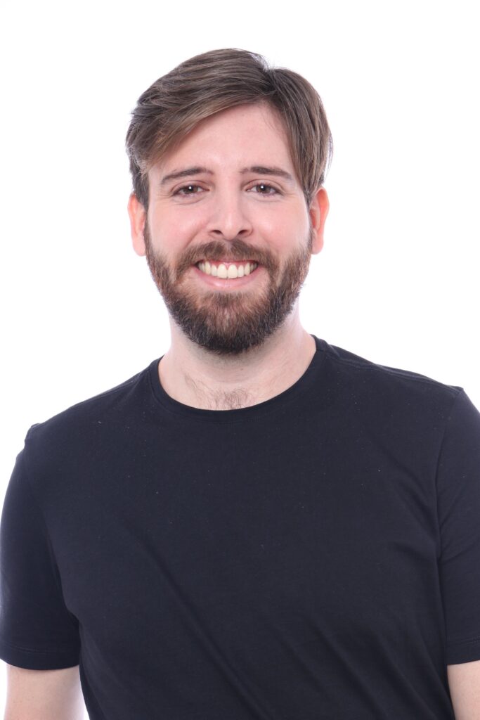 A imagem mostra um retrato de estúdio de um homem contra um fundo branco liso. Ele está centralizado e olhando diretamente para a câmera. O homem tem pele clara, cabelo castanho arrumado de forma casual e uma barba cheia. Está sorrindo amplamente, mostrando os dentes, e seus olhos estão voltados para a frente. Ele veste uma camiseta preta simples. A iluminação é clara e uniforme, típica de um estúdio fotográfico, destacando bem o rosto e criando poucas sombras. A foto é focada no rosto do homem, com o fundo branco desfocado, o que ajuda a enfatizar o sujeito. A composição é um plano médio, mostrando a cabeça e os ombros. A imagem transmite uma sensação de simplicidade e profissionalismo.