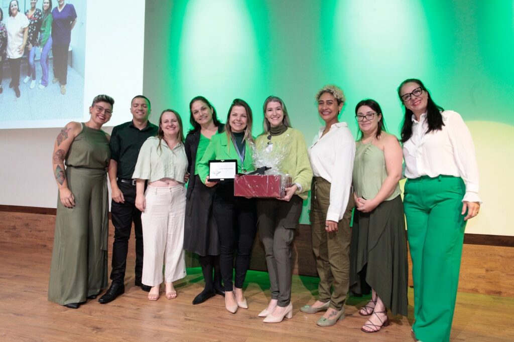 A imagem mostra um grupo de nove pessoas posando para uma foto em um palco durante um evento. O grupo é composto por oito mulheres e um homem, todos sorrindo e olhando para a câmera. A maioria está vestida de forma profissional, com roupas em tons de verde, branco e cores neutras. No centro, uma mulher segura um cesto de presente e uma placa, sugerindo que ela foi homenageada ou premiada. Ao fundo, há uma grande tela de projeção exibindo uma foto menor de quatro mulheres, e a parede atrás da tela está iluminada com luzes verdes. O ambiente parece ser formal e festivo, com iluminação clara e piso de madeira clara. A atmosfera geral transmite um momento de celebração e reconhecimento.