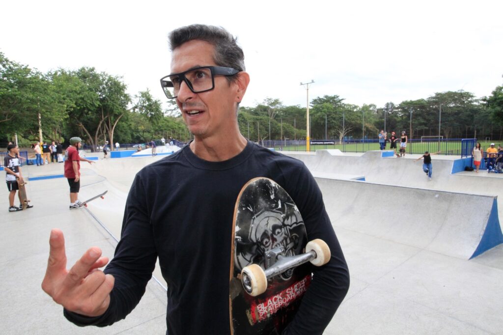 A imagem mostra um homem de meia-idade com cabelo grisalho e óculos grossos de armação preta. Ele veste uma camisa preta de manga longa e segura um skate debaixo do braço, que tem um desenho de caveira e a inscrição "BLACK SHEEP SKATEBOARDING". O homem está em um parque de skate ao ar livre, com rampas e áreas para manobras de skate ao fundo. Algumas pessoas aparecem ao fundo, algumas paradas e outras andando de skate. A cena acontece durante o dia, com uma iluminação natural e céu nublado. O ambiente parece espaçoso, com árvores e uma cerca visível ao longe. O foco principal da imagem está no homem com o skate.