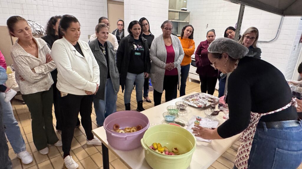 A imagem mostra um grupo de mulheres em um ambiente de cozinha profissional ou sala de aula, participando de uma oficina de culinária ou confeitaria. * No centro, uma instrutora, usando uma touca de proteção e avental, demonstra uma técnica de decoração para um grupo atento de alunas. * Sobre a mesa, há dois recipientes grandes (um lilás e um verde-claro) contendo pequenos doces ou biscoitos, além de tigelas menores com coberturas coloridas e uma bandeja com itens já preparados. * As alunas estão posicionadas ao redor da mesa, observando a demonstração com interesse. Elas vestem roupas casuais, variando entre casacos, blusas de frio e calças, o que sugere um ambiente de trabalho ou aprendizado prático. * O local possui paredes revestidas de azulejos brancos e piso de cerâmica, com uma estrutura que lembra uma cozinha industrial, reforçando o caráter educativo e profissional da cena.