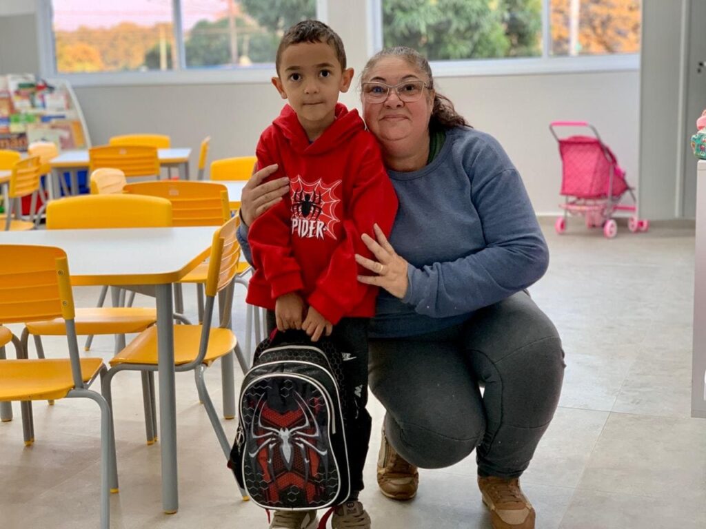 Esta imagem mostra uma cena carinhosa em um ambiente que parece ser uma sala de aula de educação infantil ou creche. * No centro, um menino pequeno está em pé, vestindo um moletom vermelho com o tema do Homem-Aranha e segurando uma mochila escolar também estampada com o mesmo personagem. Ele olha diretamente para a câmera com uma expressão serena. * Ao lado dele, uma mulher adulta está agachada, abraçando-o gentilmente. Ela usa óculos, uma blusa azul de manga longa e calças cinzas, sorrindo de forma acolhedora para a foto. * O cenário ao redor é composto por mesas e cadeiras amarelas, típicas de mobiliário escolar infantil. Ao fundo, é possível ver uma estante com livros e um carrinho de boneca cor-de-rosa, indicando um espaço de aprendizado e brincadeiras. A iluminação é clara e natural, vinda de janelas amplas atrás deles.