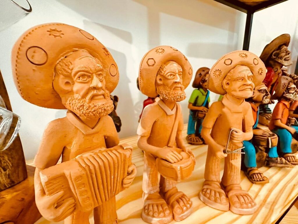 A imagem mostra várias esculturas de argila representando músicos em um cenário artesanal. As peças são feitas em tons terrosos, com detalhes que destacam instrumentos musicais como acordeão, tambor e triângulo. Os personagens usam chapéus largos e roupas simples, típicas de um estilo rural ou tradicional. As expressões faciais são sérias e concentradas, transmitindo a ideia de uma apresentação musical. As esculturas estão dispostas sobre uma superfície de madeira, com um fundo branco que mantém o foco nos detalhes das figuras. A composição sugere um grupo musical típico de uma cultura popular, evidenciando habilidade artística na modelagem da argila e na representação dos músicos em ação.