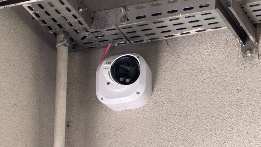A imagem mostra uma câmera de segurança branca instalada em uma parede cinza clara com textura. A câmera possui um design moderno, com uma base quadrada arredondada e uma lente preta voltada ligeiramente para baixo. Um cabo rosa está conectado à parte superior da câmera. Acima dela, há uma estrutura metálica fixada ao teto, provavelmente para gerenciamento de cabos. Ao lado esquerdo da câmera, há um tubo vertical preso à parede. A iluminação é suave, destacando os detalhes da câmera e da parede. A câmera tem a marca "CP PLUS" visível.