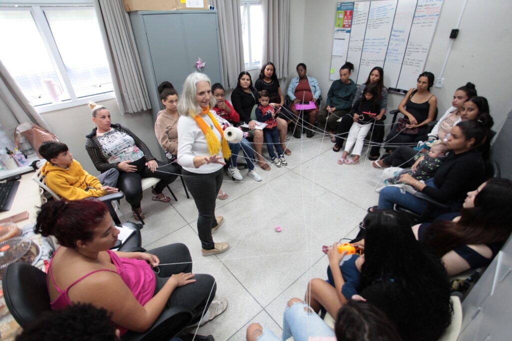 A imagem mostra um grupo de pessoas, principalmente mulheres e crianças, sentadas em círculo em uma sala clara e espaçosa, participando de uma atividade coletiva. No centro do círculo, uma mulher mais velha, com cabelo branco, usando um suéter branco e um lenço amarelo vibrante, está em pé segurando um novelo de lã branca. Ela parece estar conduzindo a atividade, gesticulando com as mãos enquanto os demais participantes seguram fios da lã, que se entrelaçam formando uma teia que conecta todos no círculo. O ambiente parece ser uma sala comunitária ou de aula, com um quadro branco ao fundo, uma estante metálica e janelas grandes com cortinas. A atmosfera transmite colaboração, interação e atenção entre os presentes. A foto foi tirada de um ângulo elevado, capturando toda a roda e a rede de fios que os une.