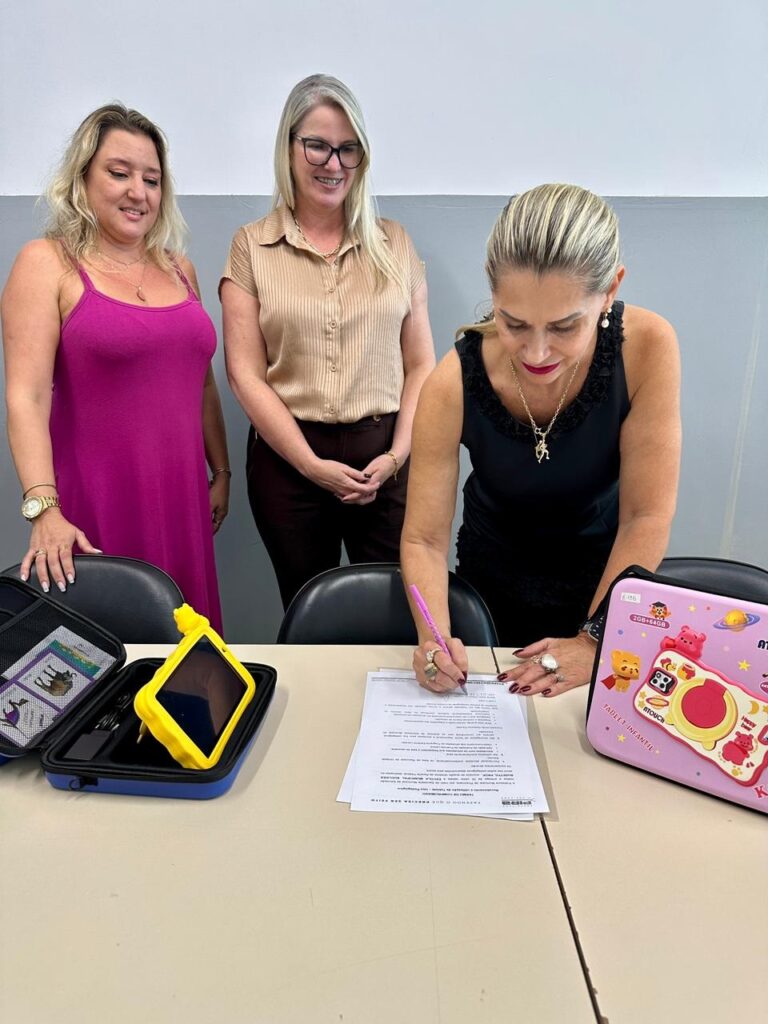 A imagem mostra três mulheres em um ambiente interno, possivelmente um escritório ou sala de reunião. A mulher em primeiro plano, com cabelo loiro preso, está assinando um documento sobre uma mesa. Ela usa uma blusa preta sem mangas e alguns acessórios, como colar e anéis. Ao fundo, duas mulheres observam atentamente: uma veste uma regata rosa e a outra uma camisa bege com calça marrom. Sobre a mesa, há dois tablets infantis em capas protetoras coloridas — um amarelo aberto e outro rosa fechado, decorado com desenhos infantis e a inscrição "TABLET INFANTIL". O documento que está sendo assinado parece ser um termo de compromisso, sugerindo que o momento é formal, possivelmente relacionado à entrega ou uso dos tablets. A iluminação é clara e o ambiente é simples, com paredes claras ao fundo.