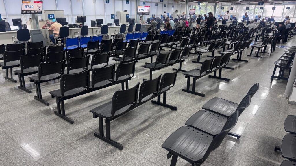 A imagem mostra uma ampla sala de espera, provavelmente de um órgão público ou banco, organizada com várias fileiras de cadeiras pretas de plástico. O ambiente é iluminado e possui um piso de granito claro. * No fundo, há uma série de guichês de atendimento com funcionários trabalhando em computadores. * Acima dos guichês, painéis eletrônicos exibem senhas, com o número "B749" em destaque no guichê 12. * O espaço está majoritariamente vazio, com poucas pessoas sentadas ou em pé próximas aos balcões de atendimento. * O ambiente transmite uma sensação de organização e funcionalidade, típica de locais de prestação de serviços ao público.