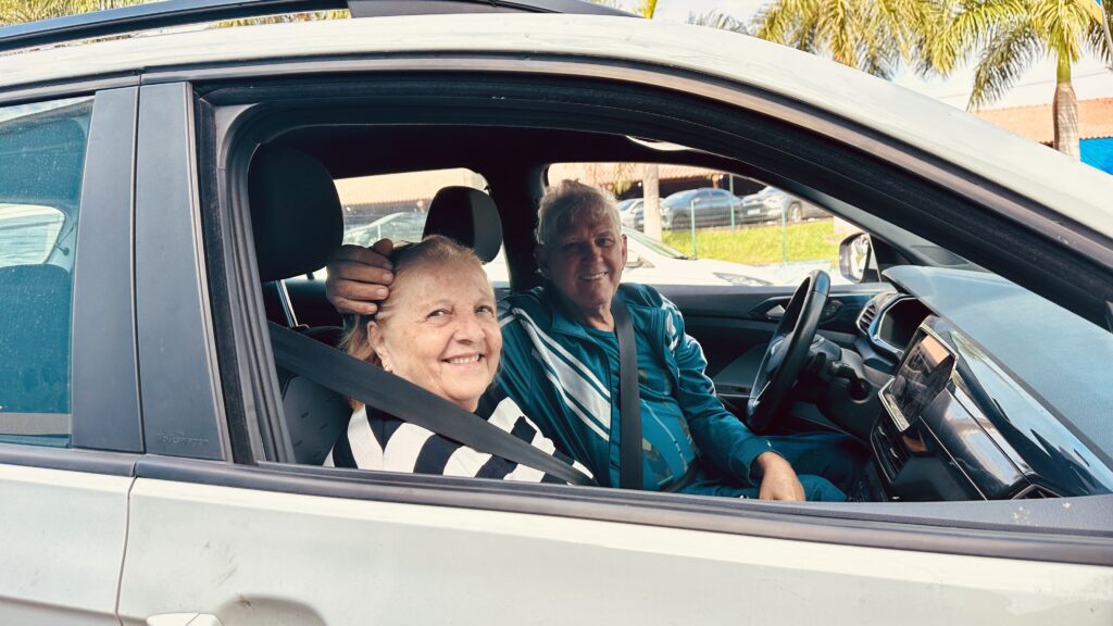 A imagem registra um momento carinhoso de um casal de idosos dentro de um carro. * O homem, que está no banco do motorista, veste uma jaqueta esportiva azul com listras brancas e mantém um sorriso aberto, enquanto coloca a mão sobre a cabeça da mulher, em um gesto de afeto. * A mulher, sentada no banco do passageiro, também sorri para a câmera, vestindo uma blusa listrada em preto e branco. * Ambos estão utilizando o cinto de segurança. O interior do veículo, incluindo o volante e o painel, é visível, e através da janela nota-se um ambiente externo iluminado, com vegetação e outros carros ao fundo, sugerindo um dia ensolarado.