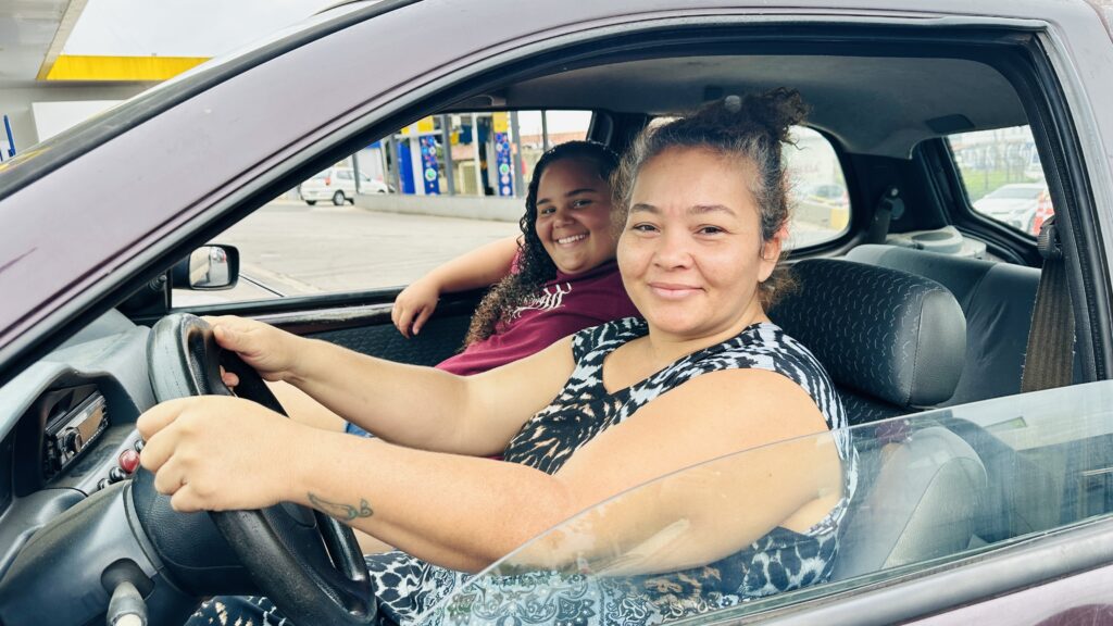 A imagem mostra duas mulheres dentro de um carro, possivelmente em um posto de combustível. A mulher no banco do motorista tem a pele clara, cabelo preso em um coque bagunçado, usa uma blusa sem mangas com estampa que lembra pele de animal e sorri de forma suave. Ela também tem uma pequena tatuagem visível no antebraço direito. A mulher no banco do passageiro, ao fundo, tem cabelo longo e cacheado, veste uma blusa vinho e sorri amplamente para a câmera. O interior do carro é escuro, com bancos cinza e volante preto. Ao fundo, pelo vidro, é possível ver parte de um posto de gasolina, com colunas amarelas e bombas de combustível azuis e brancas. A atmosfera da foto é descontraída e acolhedora, com um clima natural e iluminação suave.