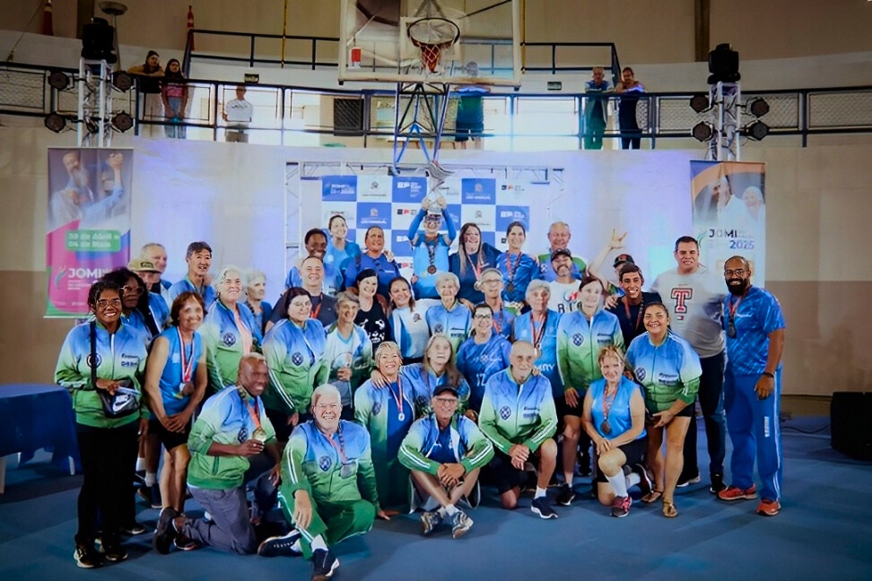A imagem mostra um grande grupo de pessoas, majoritariamente idosos, reunidos em uma quadra poliesportiva para uma cerimônia de premiação, provavelmente relacionada aos Jogos da Melhor Idade (JOMI). * O grupo está posicionado em frente a um painel com logotipos, vestindo uniformes esportivos nas cores azul e verde. Muitos dos participantes exibem medalhas penduradas no pescoço, indicando que são atletas medalhistas. * No centro, uma pessoa está erguendo um troféu acima da cabeça, celebrando a conquista. O ambiente é um ginásio iluminado, e há pessoas observando a cena de cima, em uma arquibancada ao fundo. * A atmosfera é de celebração, união e orgulho, com os participantes sorrindo e posando para a foto oficial do evento. À esquerda e à direita, banners verticais com a marca "JOMI" reforçam o contexto da competição esportiva.